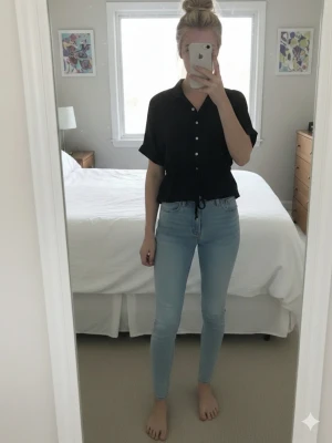 Svart blus med knappar och dragsko - Svart blus med korta ärmar, knappar framtill och dragsko i midjan. Avslappnad passform och klassisk krage. Perfekt till jeans eller kjol för en stilren look.