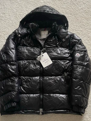 Svart dunjacka från Moncler med huva (Moncler maya)  - Snygg svart dunjacka från Moncler med glansig finish och huva. Jackan har dragkedja framtill, två sidofickor och Moncler-logga på ärmen. Perfekt för kalla vinterdagar och riktigt trendig look. Moncler maya passar till både vardagen och mer formella tillfällen. Har aldrig använts och skickas samma dag vid köp! Skriv vid mer frågor eller funderingar🤗