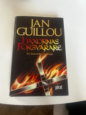 Häxornas försvarare av Jan Guillou - En svart, inbunden bok med titeln 'Häxornas försvarare' av Jan Guillou. Omslaget har eldflammor och ett kors, samt text i guld, rött och gult. Boken är ett historiskt reportage om häxprocesserna i Sverige.