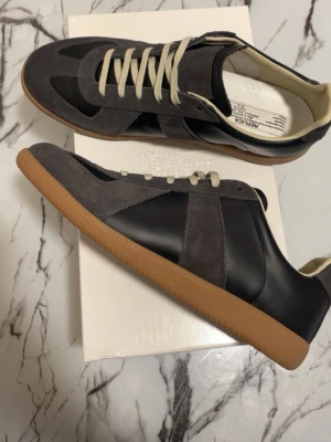 Maison margiela skor  - Maison Margiela Replica sneakers i grå mocka och svart läder med beigea skosnören och klassisk gummisula. Snygg retrostil med låg profil och vita detaljer på insidan. Perfekta för dig som gillar minimalistisk och stilren design.