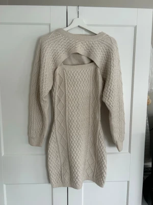 Beige stickad klänning med cut out - Trendig beige stickad klänning med snygg cut out-detalj på ryggen. Klänningen har en tillhörande bolero med ribbade muddar och ett klassiskt flätstickat mönster över hela plagget. Perfekt för dig som vill ha en mysig men ändå stilren look.