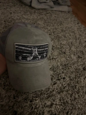 Grå keps från GOATWEAR med tryck - Säljer en grå keps från GOATWEAR med mesh på sidorna och ett svartvitt tryck framtill som visar en fotbollsspelare i aktion. Kepsen har böjd skärm och justerbar baksida. Perfekt för dig som gillar sportig stil.