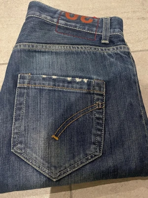Blå slitna jeans från Dondup - Säljer ett par blå jeans från Dondup med snygga slitningar och detaljerad bakficka. Jeansen har en klassisk femficksmodell och raka ben. Materialet är denim med coola slitningar och tvättade effekter för en trendig look.
