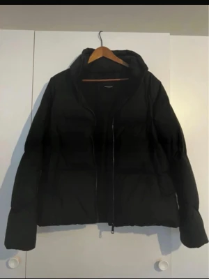 Svart dunjacka från Selected Femme  - Säljer en svart pufferjacka från SF med dragkedja framtill och hög krage. Jackan har en klassisk boxig passform och är vadderad för extra värme. Perfekt för kalla vinterdagar och enkel att matcha med olika outfits.