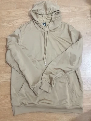 Beige hoodie med dragsko - Snygg beige hoodie med huva och dragsko. Hoodien har en klassisk passform med ribbade muddar vid ärmslut och nederkant samt en stor magficka framtill. Perfekt för en avslappnad och stilren look.