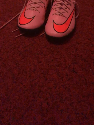 Nike fotbollsskor med rosa swoosh - Säljer ett par snygga fotbollsskor från Nike med vit bas och stora, neonrosa swoosh-loggor på ovansidan. Skorna har snörning och en greppvänlig sula för bra fäste på planen. Perfekta för dig som vill sticka ut på matchen!