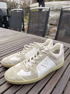 Louis Vuitton vita sneakers med mockadetaljer - Snygga vita sneakers från Louis Vuitton med beige mockadetaljer och klassisk LV-logga på sidan. Skorna har låga skaft, vita snören och en ljus gummisula. Perfekta för dig som gillar exklusiv och stilren design.