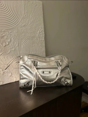 Silver metallic handväska Balenciaga - Säljer en snygg silver metallic handväska med flätade handtag och coola nitar. Väskan har en klassisk city-modell med dragkedja och detaljer i silver. Perfekt för dig som vill sticka ut med en trendig accessoar.