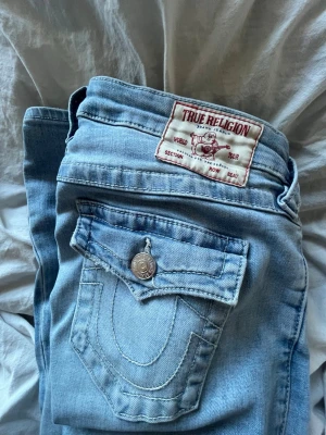 Ljusblå True Religion jeans - Säljer ett par ljusblå jeans från True Religion med klassiska kontrastsömmar och ikonisk fickdesign. Jeansen har silverfärgade knappar och detaljer samt en tydlig logga på bakfickan. Perfekta för en avslappnad och trendig look.