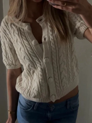 Kortärmad kofta  - Kortärmad kofta i beige med stora matchande knappar framtill. Koftan har rund halsringning och ribbade kanter. Perfekt att styla med jeans eller kjol för en trendig look.