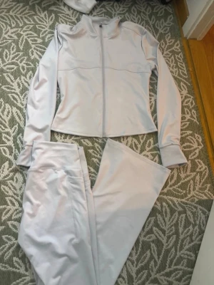 Ljusgrått träningsset med zip - Säljer ett ljusgrått träningsset med långärmad zip-jacka och matchande byxor. Jackan har hög krage och hel dragkedja framtill. Materialet är stretchigt och mjukt, perfekt för gymmet eller löprundan. Snygg och enkel design utan synliga loggor.