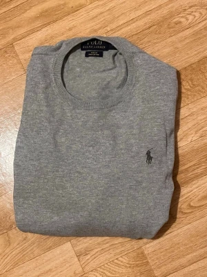 Grå stickad tröja från Polo Ralph Lauren - Klassisk grå stickad tröja från Polo Ralph Lauren med rund halsringning och diskret broderad logga på bröstet. Tröjan har långa ärmar och ribbade muddar vid ärmslut och nederkant. Perfekt till lager på lager och enkel att matcha. Mycket bra skick utan några defekter alls! Nypris: Ca 2000kr. Skriv vid minsta frågor/intresse så svarar jag så snabbt jag kan!👍