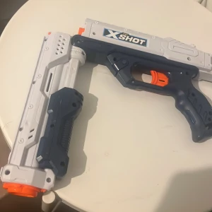 X-Shot leksakspistol i plast - Säljer en X-Shot leksakspistol i vit, mörkblå och orange plast. Pistolen har en futuristisk design med detaljerad greppyta och tydlig avtryckare. Perfekt för lek och roliga dueller med vänner.
