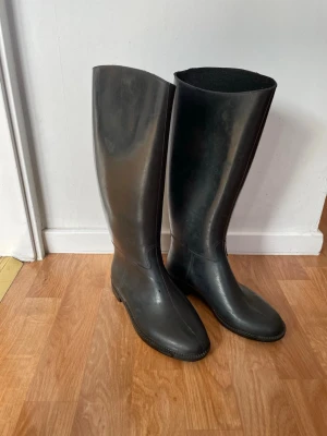 Svarta höga gummistövlar - Säljer ett par svarta höga gummistövlar med klassisk, stilren design. Stövlarna är tillverkade i blankt gummi och har rundad tå samt låg klack. Perfekta för regniga dagar och outdoor-looks.