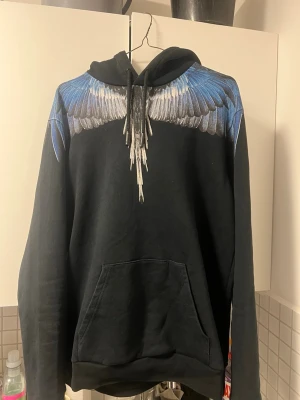 Äkta Marcelo Burlon hoodie  - Snygg svart hoodie med ett unikt tryck av blå och vita vingar över axlarna och ner över bröstet. Hoodien har huva med snören och en stor magficka framtill. Perfekt för dig som vill sticka ut med en cool och kreativ design.