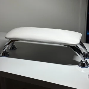 Vitt armstöd, till naglar  - Nagelbordsstöd i stilren vit färg med mjuk, rektangulär dyna och stabila kromade metallben. Perfekt för att vila händerna vid nagelbehandlingar. Dynan är klädd i konstläder och ger både komfort och stöd under manikyr.