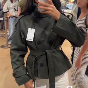 Kort mörkgrön trenchcoat från Bershka - Stilren mörkgrön trenchcoat från Bershka med dubbelknäppning, axelklaffar och knytskärp i midjan. Jackan har hög krage, långa ärmar och två sidofickor. Perfekt för dig som vill ha en trendig och kortare trenchcoat i bomull.