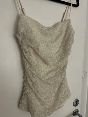 Vit spets linnetopp med tunna band - Söt vit linnetopp med tunna axelband och vacker spets över hela toppen. Draperad framtill för en snygg och trendig look. Perfekt att styla med jeans eller kjol för en fräsch outfit.