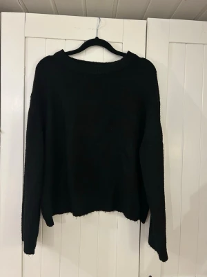 Svart stickad tröja från 157 - En enkel och stilren svart stickad tröja från 157 med rund halsringning och långa ärmar. Tröjan har en rak passform och ribbstickade muddar vid hals, ärmslut och nederkant. Perfekt att matcha med jeans eller kjol.