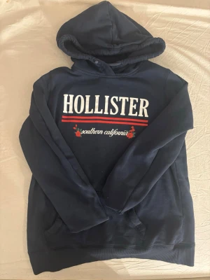 Mörkblå hoodie från Hollister - Snygg mörkblå hoodie från Hollister med stor vit logga och röda detaljer på bröstet. Hoodien har huva och klassisk känguruficka framtill. Materialet är mjukt och bekvämt, perfekt för chill dagar. Dekorativa blommor och texten 'southern california' ger en extra touch.