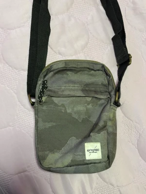 Grå camo axelväska Springfield - Snygg axelväska i grått camomönster från Springfield. Väskan har justerbar svart axelrem, dragkedja upptill och en praktisk ytterficka med dragkedja. Tillverkad i slitstarkt tyg och har en liten vit logolapp framtill.