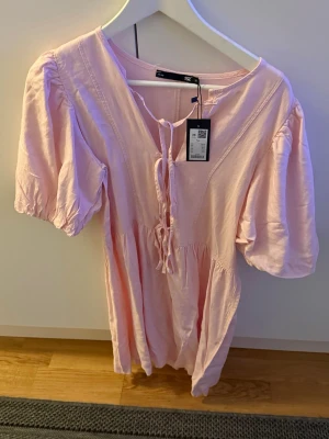 Ljusrosa kort klänning med snörning - Supersöt ljusrosa kort klänning från Rosie med puffärmar och snörning framtill. Klänningen har en avslappnad passform och är gjord i ett mjukt bomullsmaterial. Perfekt för varma dagar och ger en romantisk vibe.