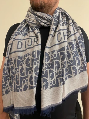 Gråblå Dior halsduk med logotypmönster - Snygg halsduk från Dior i gråblått med tydligt logotypmönster och texten 'DIOR' invävd. Halsduken har fransade kanter och är tillverkad i ett mjukt tyg som känns lyxigt och bekvämt runt halsen. Perfekt accessoar för att lyfta din outfit.