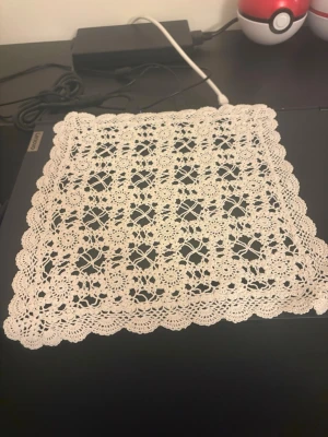 Virkad vit duk i spetsmönster - Säljer en handgjord, fyrkantig virkad duk i vitt med detaljerat spetsmönster och vågig kant. Perfekt som dekoration på bord eller hylla. Materialet är tunt garn, troligen bomull.