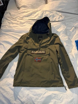 Olivgrön vindjacka från Napapijri - Snygg olivgrön vindjacka från Napapijri med stor ficka framtill, dragkedja vid halsen och huva. Materialet är slitstarkt och vattenavvisande, perfekt för blåsiga dagar.