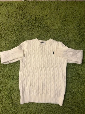 Vit kabelstickad tröja Polo Ralph Lauren - Kabelstickad vit tröja från Polo Ralph Lauren med rund halsringning och svart broderad logga på bröstet. Tröjan har långa ärmar och ribbade muddar vid ärmslut och nederkant. Klassisk och tidlös design i mjukt material.