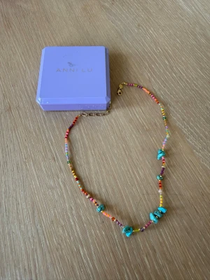 Färgglatt pärlhalsband från Anni Lu - Unikt halsband från Anni Lu med små färgglada pärlor i olika nyanser och turkosa stenar. Halsbandet har en tunn guldfärgad kedja och lås. Perfekt för dig som gillar lekfulla accessoarer med boho-vibe.