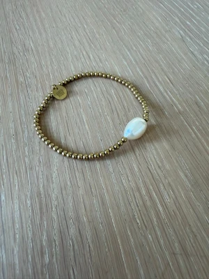Guldfärgat armband med pärldetalj - Snyggt armband med små guldfärgade kulor och en större vit pärla som detalj. Armbandet har en enkel och stilren design med en liten rund bricka nära låset. Perfekt för dig som gillar minimalistiska accessoarer.