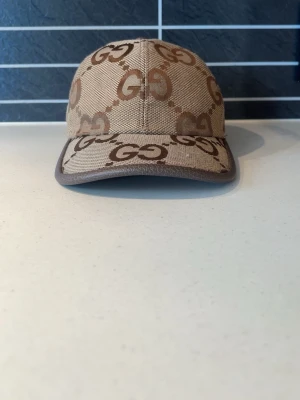 Beige keps med GG-mönster  - Snygg beige keps med det klassiska GG-monogrammet i brunt över hela kepsen. Kepsen har böjd skärm och bruna kanter. Tillverkad i canvasmaterial som ger en exklusiv känsla och passar perfekt till streetwear-stilen.