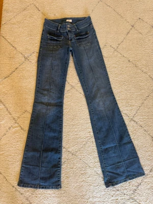 Blå utsvängda jeans från Nelly - Säljer ett par blå jeans från Nelly med utsvängda ben och dubbla knappar i midjan. Jeansen har två stora fickor framtill med lock och dekorativa sömmar längs benen. Tillverkade i klassisk denim med en cool vintagekänsla.