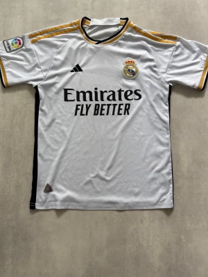 Real Madrid 23/24 matchtröja Adidas vit - Snygg vit Real Madrid matchtröja från Adidas med svarta och guldiga detaljer. Tröjan har klubbmärke, Adidas-logga och sponsortryck på bröstet samt La Liga-märke på ärmen. Tillverkad i lätt och ventilerande material, perfekt för fotboll, träning eller sommaren, som ger en trendig och en unik fotbollslook.
