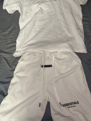 Vitt set från Essentials Fear of God - Säljer ett vitt set från Essentials Fear of God med t-shirt och shorts. Båda plaggen har minimalistisk design med logga tryckt på bröstet och benet. Setet har en avslappnad passform och är gjort i mjukt material, perfekt för en clean och stilren look. Pris kan diskuteras vid snabb affär.