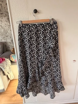 Svart blommig långkjol med volang - Säljer en svart långkjol med vita blommor och volangkant. Kjolen har en flowy passform och är gjord i ett lätt, tunt material som känns luftigt. 