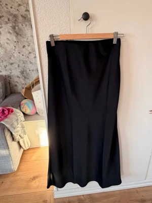 Svart långkjol  - Stilren svart långkjol i glansigt satinliknande material. Kjolen har en rak passform och faller snyggt längs benen. 