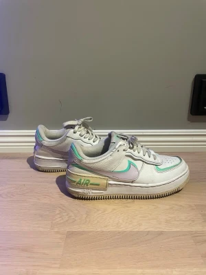Vita Nike Air Force 1 Shadow sneakers - Nike Air Force 1 Shadow sneakers i vitt läder med detaljer i mintgrönt och ljuslila. Skorna har en chunky sula, perforerad tå och dubbla lager på swoosh-loggan och hälen. Klassisk låg modell med snörning och retrovibbar.