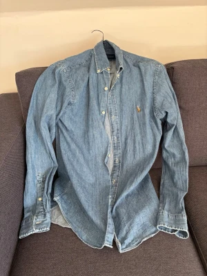 Blå jeansskjorta från Polo Ralph Lauren - Säljer en klassisk blå jeansskjorta från Polo Ralph Lauren med knappstängning och lång ärm. Skjortan har den ikoniska broderade loggan på bröstet och är tillverkad i mjuk bomull. Perfekt till jeans eller chinos för en avslappnad look. Stl M