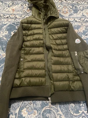 Olivgrön pufferjacka från Moncler - Säljer en olivgrön pufferjacka från Moncler med stickade ärmar och huva. Jackan har quiltad front i glansigt material, dragkedja framtill och Moncler-logga på ena ärmen. Perfekt för kalla vinterdagar och riktigt snygg streetstil.