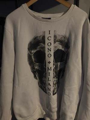 Vit sweatshirt Icono Milano dödskalle - Säljer en vit sweatshirt från Icono Milano med en stor svart dödskalle-tryck på framsidan och texten 'ICONO MILANO' vertikalt. Tröjan har rund halsringning och ribbade muddar. Perfekt för dig som gillar streetwear och grafiska prints.