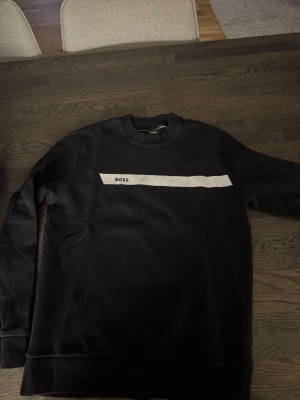 Hugo Boss sweatshirt  - Svart sweatshirt från BOSS med vit rand och logga på bröstet. Klassisk rund halsringning och långa ärmar. Tillverkad i mjukt material som känns skönt mot huden. Perfekt för en clean och stilren look.