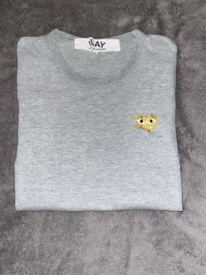 Grå t-shirt från Play Comme des Garçons - Grå t-shirt från Play Comme des Garçons med ett litet gult hjärta med ögon broderat på bröstet. Klassisk rund halsringning och korta ärmar. Perfekt för en stilren och trendig look.