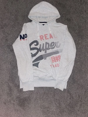 Ljusgrå hoodie från Superdry - Ljusgrå hoodie från Superdry med stor tryckt logga i marinblått och rött framtill. Huva med dragsko och ribbade muddar vid ärmar och nederkant. Tryck även på ärmen och ryggen. Perfekt för en avslappnad streetwear-look.