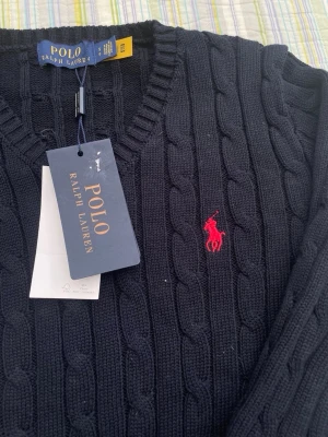 Mörkblå kabelstickad tröja Polo Ralph Lauren - Mörkblå kabelstickad tröja från Polo Ralph Lauren med klassisk röd broderad logga på bröstet. Tröjan har rund halsringning, långa ärmar och ribbstickade muddar. Perfekt för en stilren och tidlös look.