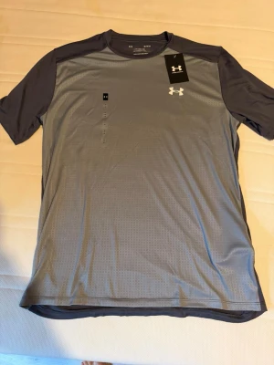 Grå tränings-tshirt från Under Armour - Snygg grå tränings-tshirt från Under Armour med mörkare grå ärmar och vitt logotryck på bröstet. Tillverkad i ett lätt och snabbtorkande funktionsmaterial som passar perfekt till gymmet eller löprundan. Klassisk rund hals och korta ärmar.