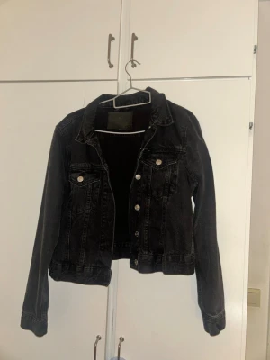 Svart jeansjacka med silverknappar - Snygg svart jeansjacka med klassisk krage och silverfärgade knappar framtill. Två bröstfickor med lock och knapp, samt raka ärmar. Perfekt att slänga över en hoodie eller t-shirt för en cool och avslappnad look.