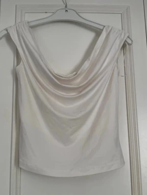Vit  topp med draperad urringning - Säljer en vit  topp med snygg draperad urringning och ärmlös design. Toppen har en mjuk, glansig yta som ger en lyxig känsla och passar perfekt till jeans eller kjol. Enkel att styla för en trendig look.