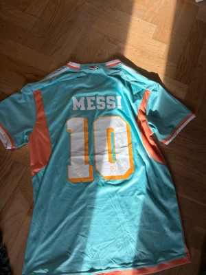 Turkos Inter Miami Messi tröja - Säljer en turkos fotbollströja med Messi och nummer 10 på ryggen. Framsidan har Inter Miami-logga och texten MIAMI. Tröjan har vita och orange detaljer längs sidor och ärmslut, samt rund hals. Perfekt för dig som gillar fotboll och Messi.